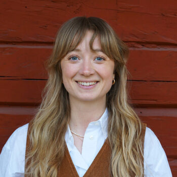 Porträtt på Johanna som står mot en rödmålad timrad vägg.